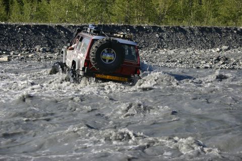 2003 Jeep TJ - A red SUV navigates a rocky riverbed, kicking up water as it drives.
