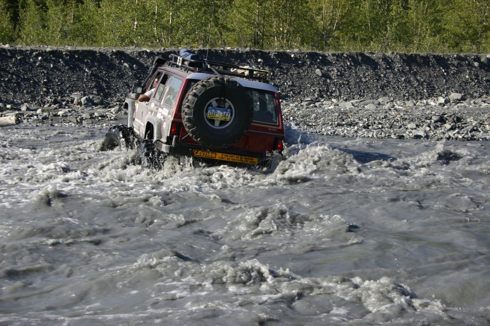 Red SUV driving through murky water, spare tire on rear, roof rack, caution sticker visible