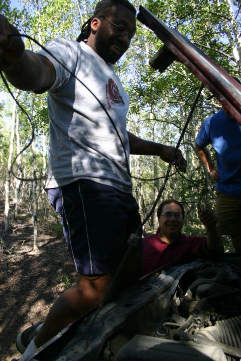 2003 Jeep TJ - People are gathered around the open engine bay of a vehicle, appearing to work on it in a wooded area.