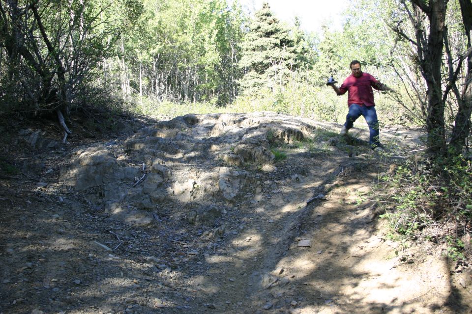 Man in red shirt and blue jeans stands on a rocky, wooded trail