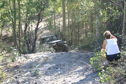 2003 Jeep TJ - A green Jeep TJ navigates a steep, rocky trail surrounded by trees, with a person observing from the side.