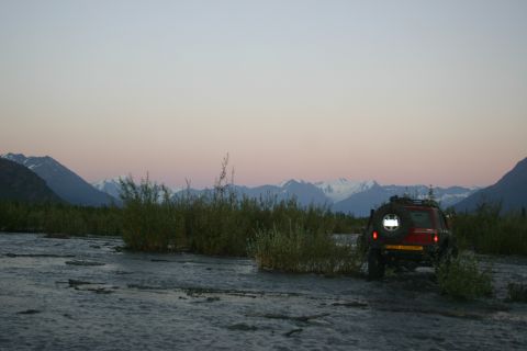2003 Jeep TJ - SUV navigates a muddy riverbed with a mountain range visible in the distance at twilight.