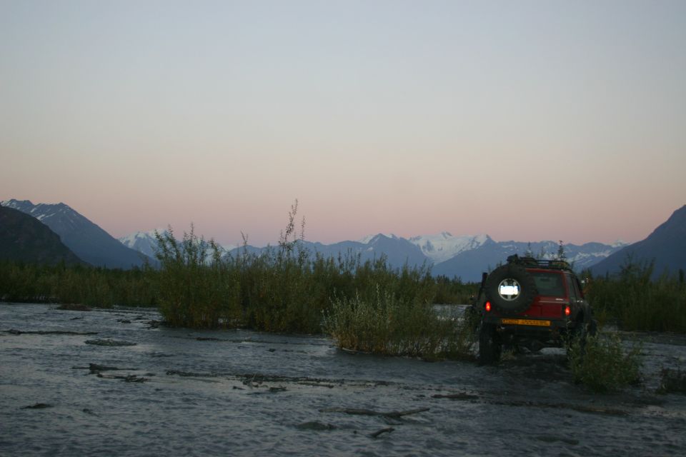 Red SUV with spare tire on back driving in a riverbed, mountains and dusk sky behind