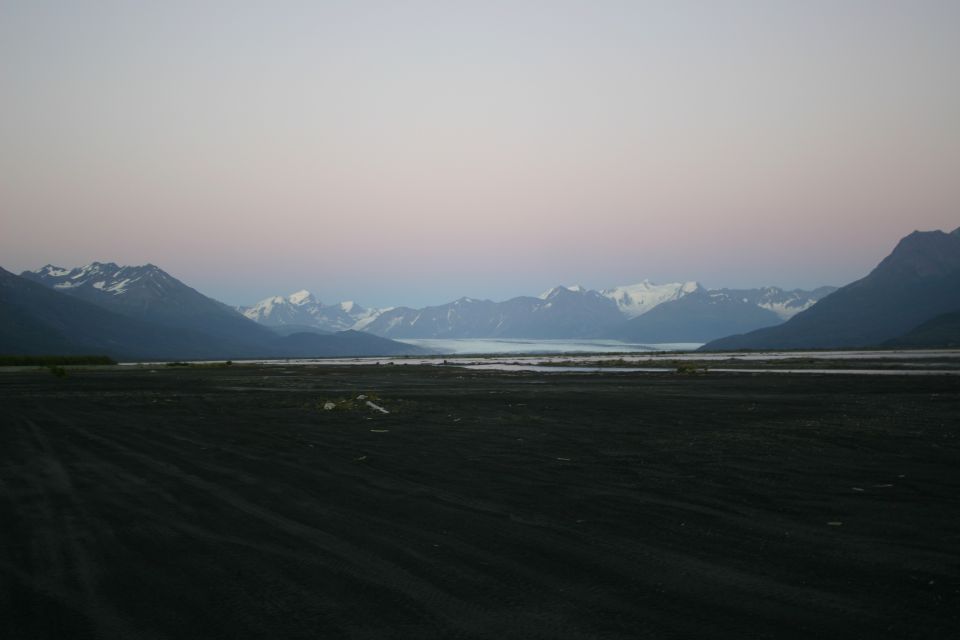 Vast dark foreground with tire tracks leads to a wide, flat valley with a river or lake, backed by immense, snow-capped mountains under a pale pink and blue sky