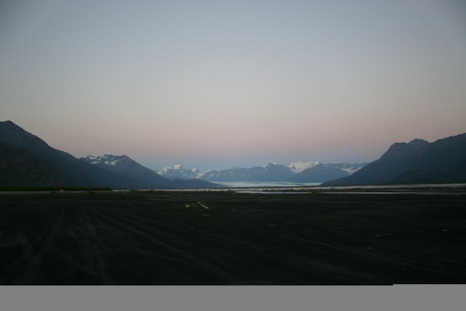 Dark, barren valley floor with tire tracks leads to a wide, calm river or lake reflecting snow-capped mountains under a pale pink and blue twilight sky