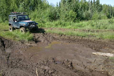 2003 Jeep TJ - A Jeep Wrangler navigates a challenging muddy off-road course with dense trees in the background.