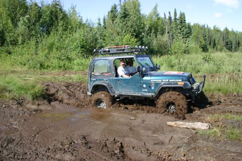 2003 Jeep TJ - A teal Jeep TJ is stuck in a muddy trail, with its tires submerged in thick mud.