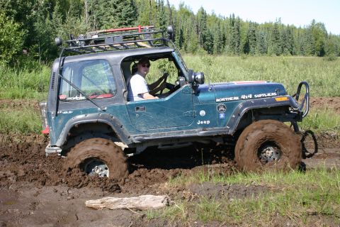 2003 Jeep TJ - A teal Jeep Wrangler TJ navigates a muddy off-road course, its front right wheel deep in the mire.
