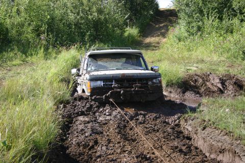 2003 Jeep TJ - A white SUV is deeply stuck in thick mud on an off-road trail, with a rope attached to its front for towing.