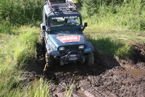 2003 Jeep TJ - A dark green Jeep TJ navigates a muddy off-road trail, its front tires deep in the mire.
