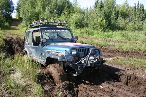 2003 Jeep TJ - A blue Jeep Wrangler TJ navigates a muddy off-road trail, its tires deep in the mire.