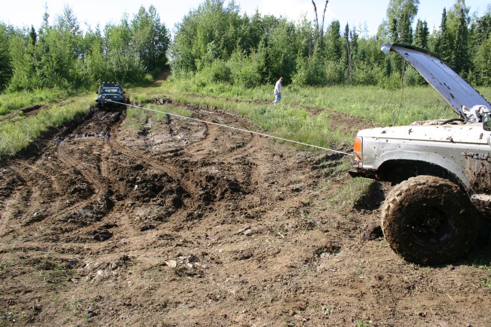 Two SUVs stuck in mud, one pulling the other with a tow rope. Towing vehicle has hood open
