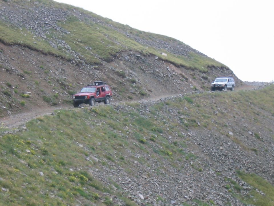 Red 1997 Jeep Cherokee and white SUV driving on a narrow dirt trail up a steep, rocky mountain