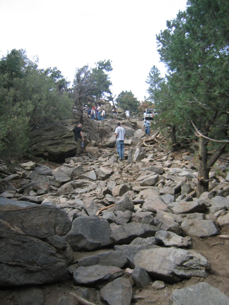 White SUVs navigating a steep, rocky mountain trail with trees and people
