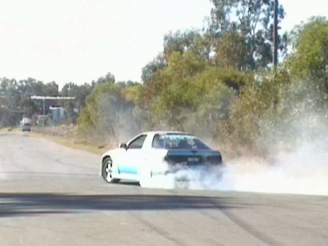 White and teal coupe doing a burnout on an open rural road, thick smoke pouring off rear tires