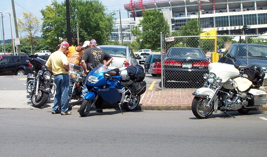 Three motorcycles parked on asphalt, with people standing around two of them. A white motorcycle is to the right