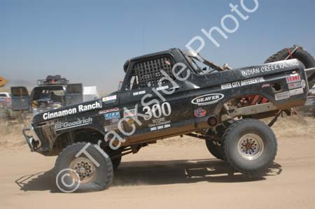 1990 Ford Bronco - black tube-chassis off-road race truck numbered 300 with sponsor decals catches air on a dirt course.