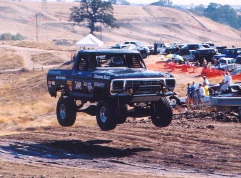 1990 Ford Bronco - black off-road race truck marked 300 launches off a dirt jump as spectators and pop-up tents line the hillside course.