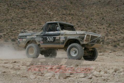 1990 Ford Bronco - black off-road race truck marked 300 kicks up dust cornering on a rocky desert course beneath barren hills.