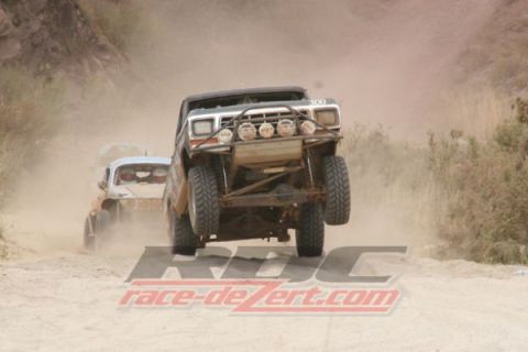 1990 Ford Bronco - black off-road race truck with round light pods jumps head-on through a dusty canyon, a second rig trailing in the dust.