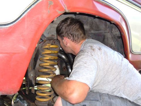 1978 Ford Bronco - A mechanic works on the suspension of a red vehicle, focusing on the yellow coil spring and exhaust headers.
