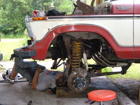 1978 Ford Bronco - A mechanic works on the underside of a red and white Ford Bronco Custom, with custom exhaust headers visible.