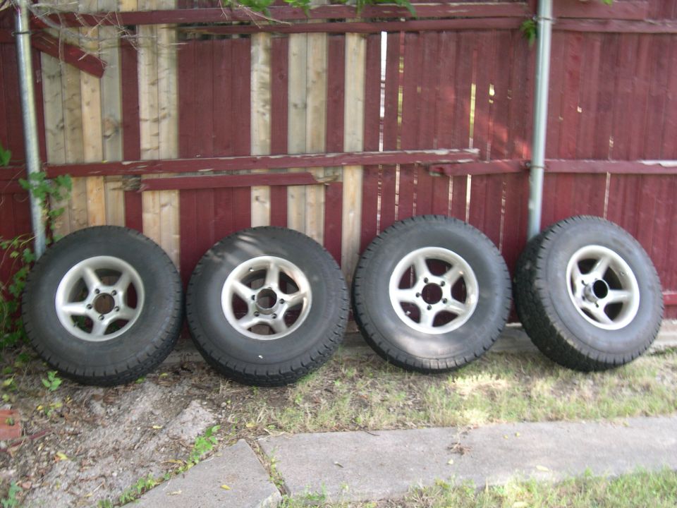 Four off-road tires on silver five-spoke alloy wheels leaning against a red wooden fence