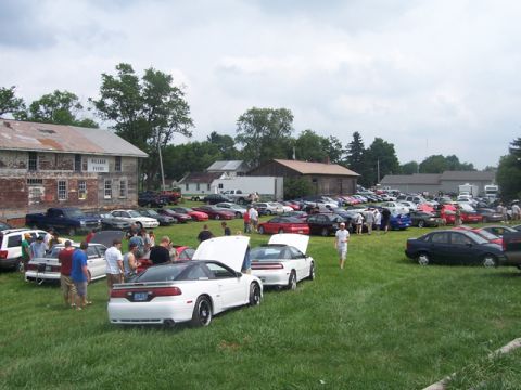 1990 Ford Probe - A gathering of cars and people on a grassy field with buildings in the background.