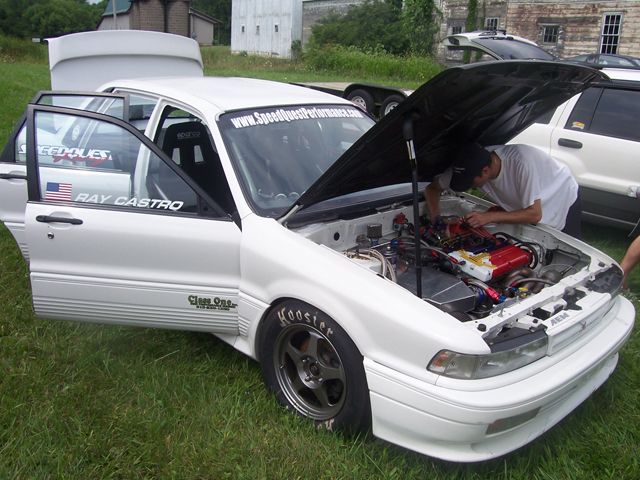 White sedan with doors open, engine being worked on, racing tires, and a "SpeedQuestPerformance.com" banner