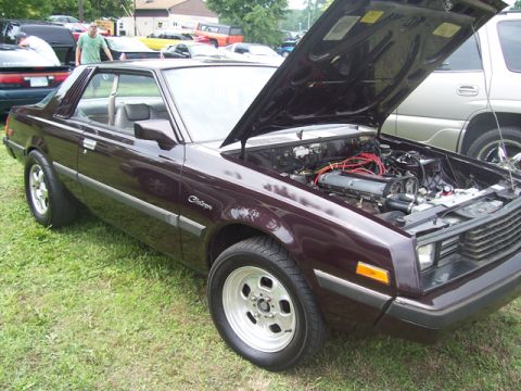 1990 Ford Probe - This maroon coupe, likely from the early 1980s, has its hood open, revealing the engine bay and other vehicles in the background.