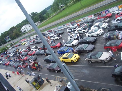 1990 Ford Probe - A large gathering of cars are parked on a wet asphalt lot, with a racetrack visible in the background.