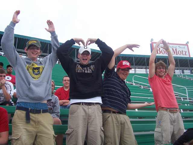 Four young men with arms raised in stadium seating, one forming a heart shape with his hands