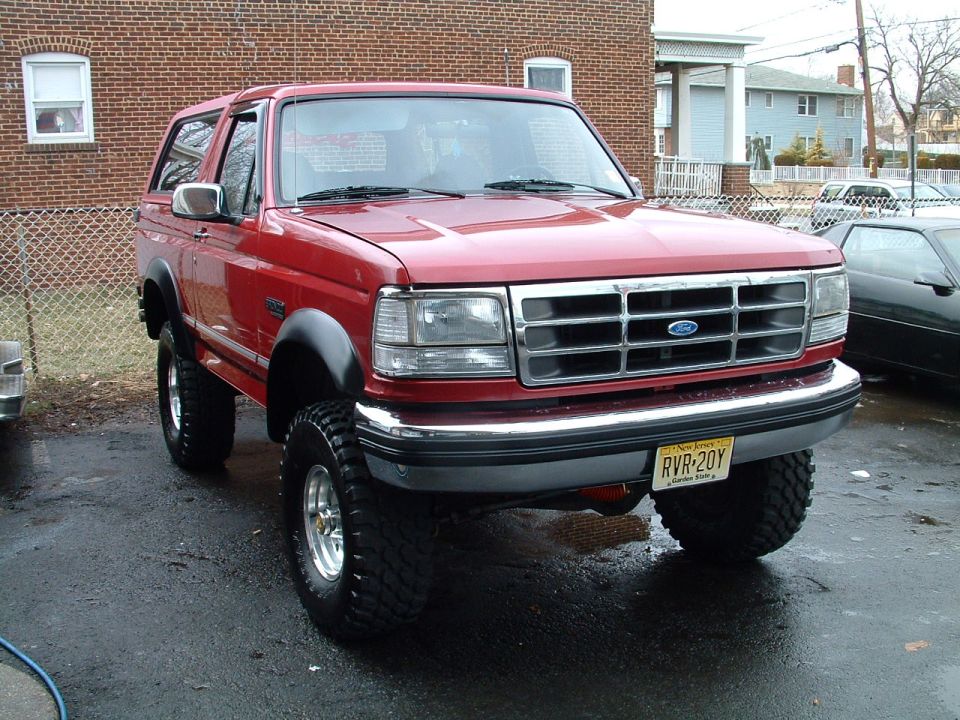 Red lifted Ford Bronco on mud-terrain tires, front 3/4 view in a wet brick-house driveway