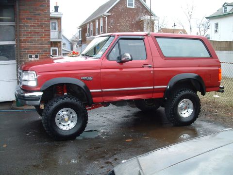 1994 Ford Bronco - Red full-size Ford Bronco with a suspension lift, oversized mud tires on chrome wheels, black fender flares and a removable hardtop, parked…