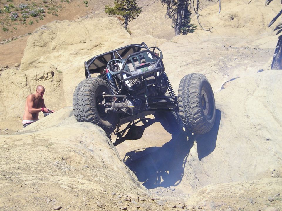Off-road buggy with a roll cage and large tires navigating a rocky incline, a man stands nearby