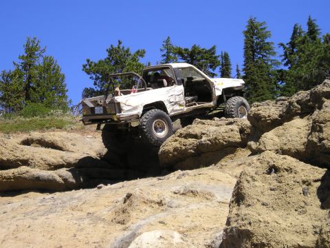 1989 Ford Bronco - White doorless mini-truck with a roll cage, tube front bumper and heavily dented body descends a tan rock ledge, shot from below against bl…