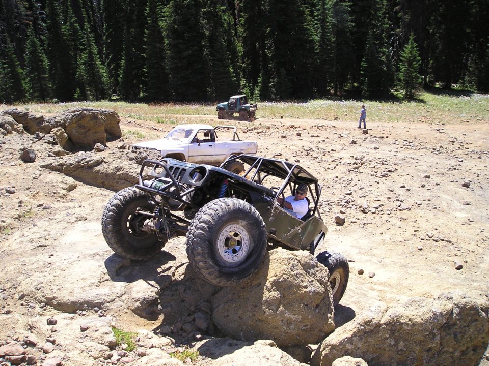 Custom buggy with roll cage climbing over a rock, white pickup and green ATV behind it
