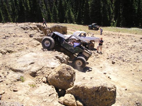1989 Ford Bronco - Elevated three-quarter view of a green tube-chassis rock buggy nosing off a rocky ledge on a dirt trail, with spectators watching and a str…