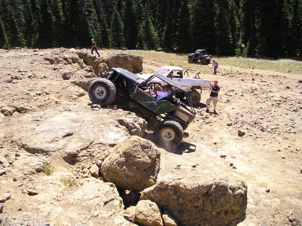 Green caged rock buggy on oversized tires dropping off a ledge with spectators and rigs behind