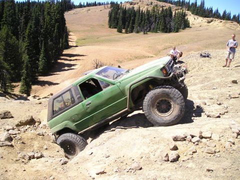 1989 Ford Bronco - An off-road SUV navigates a steep, rocky incline in a mountainous, wooded area.
