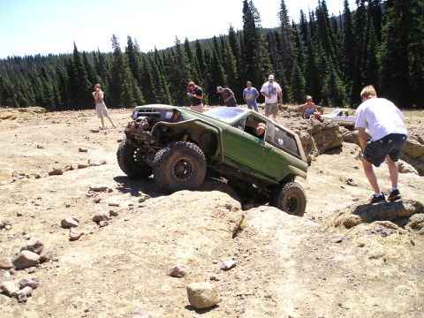 1989 Ford Bronco - Green cut-down 4x4 SUV with a chopped roof, bobbed rear and winch-equipped front bumper crawls across a rock shelf in a pine-forested alpin…