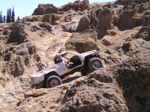 1989 Ford Bronco - Side view of a stripped white full-size 4x4 pickup-style rig climbing a steep tan volcanic rock face, doors and bed removed with an exposed…
