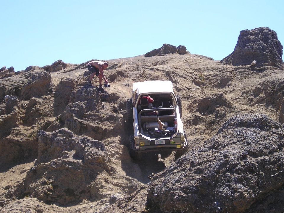 White SUV with roll cage climbing a steep, rocky hill. A person stands nearby. The vehicle's rear is visible
