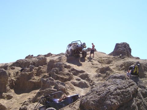 1989 Ford Bronco - A stripped white full-size 4x4 with a roll cage and no doors crawls over a light-colored rock outcrop under a clear blue sky while a spotte…