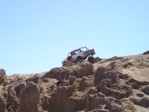 1989 Ford Bronco - Distant side view of a stripped white doorless 4x4 with an exposed roll cage perched high on a tan rock ridge against a clear blue sky. It…