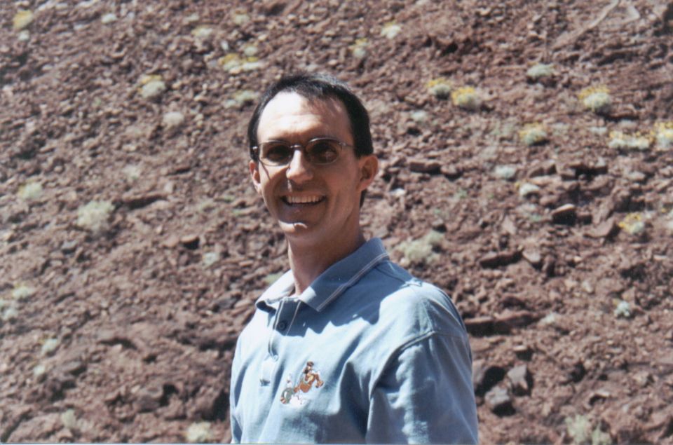 Man wearing glasses and a blue polo shirt smiles in front of a rocky, desert hillside. Watermark reads SUPER MOTORS