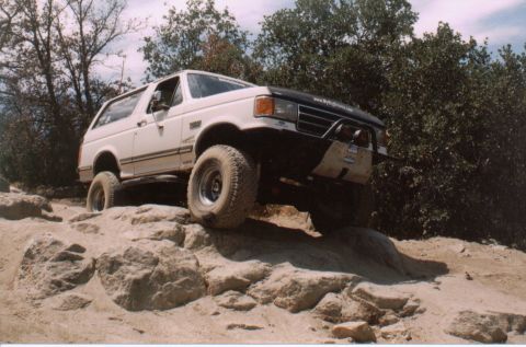 1990 Ford Bronco - White lifted Ford Bronco flexing over pale sandstone rock ledges on a wooded trail, shot low from the driver's-side front three-quarter. We…