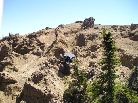 1989 Ford Bronco - A tube-framed rock crawler buggy with a light-colored roof and large off-road tires climbs a steep, rocky, tan-colored terrain, while a shi…