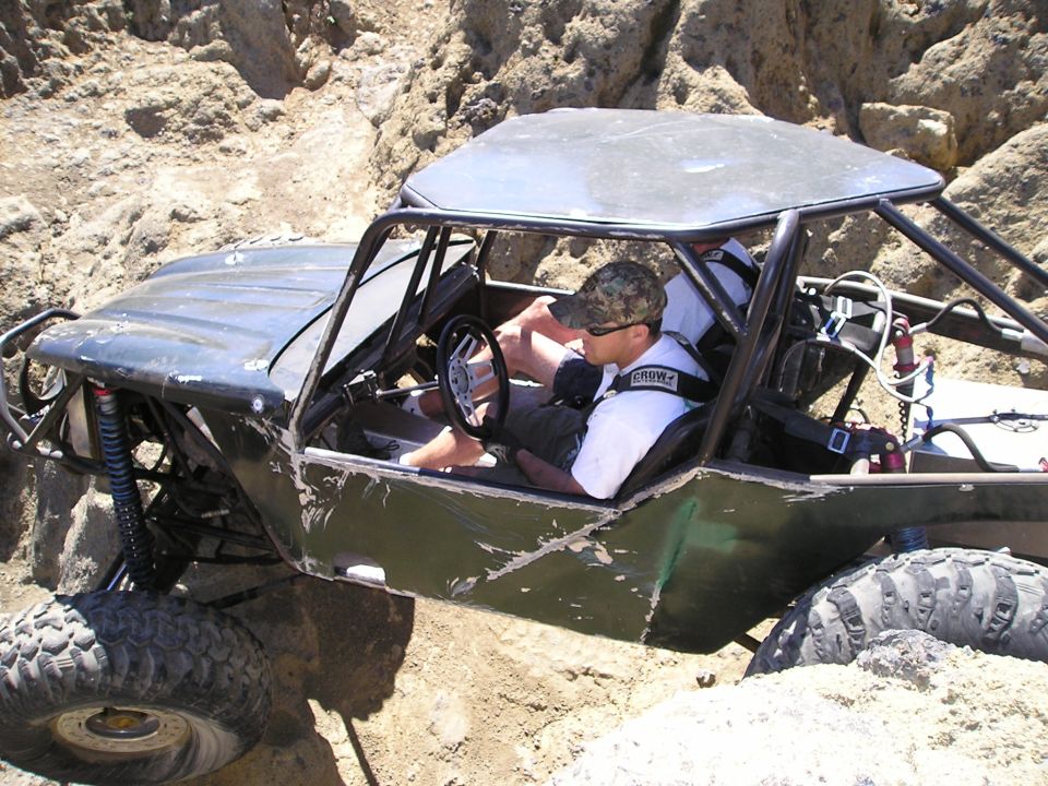 Off-road buggy with a driver and passenger navigating a rocky trail. The vehicle has a roll cage and large tires