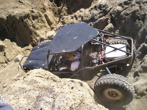 1989 Ford Bronco - Overhead three-quarter view down onto a black tube-chassis rock buggy working through a tan rock crevice, the helmet-less driver visible at…